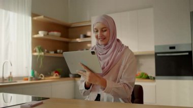 Gülümseyen Müslüman kadın, güneş ışığı mutfağında sandalyede oturan tablet bilgisayar kullanıyor. Kamera hareket ediyor. 360 derece