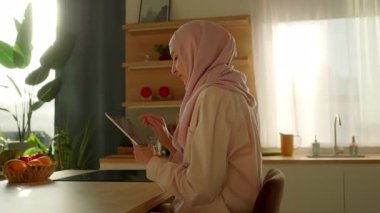 Gün ışığında güzel Müslüman kadın evdeki mutfakta tablet bilgisayar kullanıyor. Düşünüyorum. Kamera hareket ediyor. 360 derece