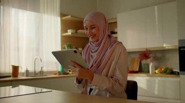 Mutlu Müslüman kadın güneş ışığı mutfağındaki masa başında tablet bilgisayar kullanıyor. Evde. Kamera hareket ediyor. 360 derece