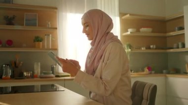 Güzel müslüman kadının manzarası güneş ışığı mutfağında oturan tablet bilgisayar kullanıyor. Kamera hareket ediyor. 360 derece