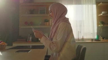 Modern mutfakta tesettürlü mutlu müslüman kadın gün ışığında gülümseyen telefonu kullanır. Kamera hareket ediyor. 360 derece