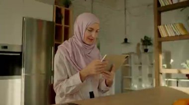 Genç ve güzel müslüman bayanın olduğu hızlandırılmış video güneş ışığı mutfağında oturan tablet bilgisayar kullanıyor. Kamera hareket ediyor. 360 derece