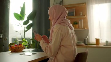 tesettürlü gülümseyen müslüman kadın, telefon mesajı yazarak sosyal medyada sohbet ediyor gün ışığında mutfakta oturuyor. Mutlu ol. Kamera hareket ediyor. 360 derece
