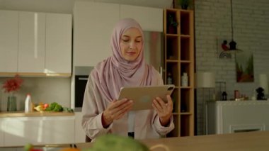 Güzel Müslüman kadının portresi güneş ışığı mutfağında oturan tablet bilgisayar kullanıyor. Kamera hareket ediyor. 360 derece