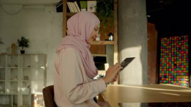 Genç ve güzel bir Müslüman kadın güneş ışığı mutfağındaki masa başında tablet bilgisayar kullanıyor. Kamera hareket ediyor. 360 derece