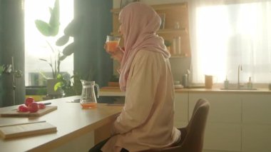 Genç tesettürlü, tesettürlü Müslüman kadın modern aydınlık mutfakta meyve suyu içiyor. Kamera hareket ediyor. 360 derece