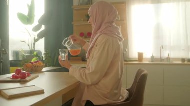 Tesettürlü, tesettürlü, gülümseyen Müslüman kadın modern günışığı mutfağında meyve suyu içiyor. Kamera hareket ediyor. 360 derece