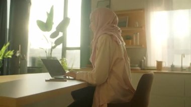 Güzel Müslüman iş kadını başörtüsü takıyor evdeki masada dizüstü bilgisayarla çalışıyor. Güneş ışığı. Kamera hareket ediyor. 360 derece