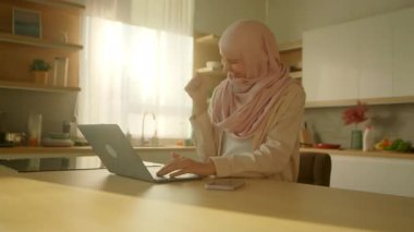 Sırıtan müslüman iş kadını dizüstü bilgisayar başında çalışırken evde mutlu oluyor. Güneş ışığı. Kamera hareket ediyor. 360 derece