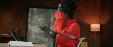 Afro saçlı, gülümseyen Afrikalı Amerikalı bir kadının 360 derecelik görüntüsü. Dijital tabletle masada otururken kulaklık takan turuncu kazaklı bir kız. Notları olan şık bir duvar
