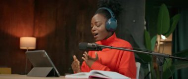 Modern stüdyoda radyo programı kaydedilirken fevkalade afro saç stiliyle mikrofona konuşan enerjik ve çekici genç Afrikalı Amerikalı kadının görüntüsü. Radyo sunucusu dijital tablet kullanıyor. Kamera.