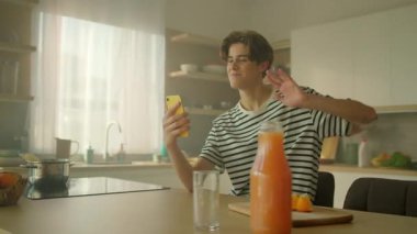 Kahvaltı masasında oturup video aracılığıyla arayan neşeli, genç, beyaz bir adamı vurmak. Güzel görünüşlü, gülümseyen bir adamın başparmağını kaldırdığı ve kameraya barış işareti yaptığı bir görüntü. Mutfağa. Parallaks