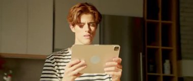 Elinde dijital tablet ve arama uygulaması olan odaklanmış genç bir kızıl adamın görüntüleri. Düşünceli adam kendi kendine konuşuyor ve mutfakta geziniyor. 360 derecelik izleme atışı. Parallaks etkisi. Döndürme görünümü