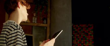 Çizgili tişörtlü, dijital tablet tutan ve ekrana dokunan rahat genç beyaz bir adam. Adam arkadaşından komik mesajlar okuyor ve gülüyor. İz sürme hareket ediyor. 360 derece mutfak. Parallaks