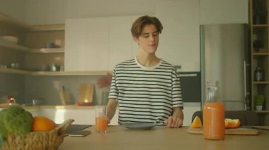 Genç beyaz adam mutfağa portakal dilimi koyuyor. Akıllı telefon kullanırken camdan portakal suyu içen adam. Yemek masası. Kamera hareket ediyor. Parlak stüdyo. Gündüz vakti. Kapalı