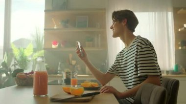 360 derece atış. Beyaz erkek öğrencinin elinde akıllı telefon tutarken ve video aramasıyla aradığı görüntüler. Mutfakta taze malzemelerle omuz silkip kahvaltı masasında konuşan mutlu adam.