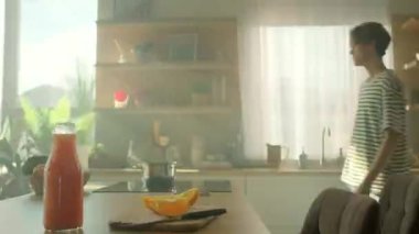 Enerjik hareketler, genç gülümseyen beyaz adamın mutfakta vakit geçirmesini takip ediyor. Akıllı telefon kullanıp portakal suyu içen neşeli bir adamın görüntüsü. Parallaks etkisi. Gündüz vakti. İletişim
