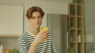 Masada oturan ve modern mutfakta akıllı telefon kullanan genç beyaz bir adamın hareketli görüntüsü. Adam ekrandaki cihazlara bakıyor ve mesajları kontrol ediyor. İz sürme hareket ediyor. Parallaks etkisi