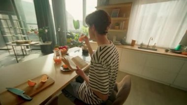 Kafkasyalı genç adam elinde kitap tutuyor ve kahvaltı masasında otururken portakal suyu içiyor. Modern rahat stüdyoda kitabı rafa koyan adam. 360 derecelik izleme atışı