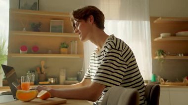 Dizüstü bilgisayarda oturan ve video aracılığıyla konuşan genç bir erkeğin görüntüsü. Evde çalışma günü. Uzak bir iş. İz sürme sahnesinde dolaşıyorum. Klavyede yazan gülümseyen beyaz adam. Stüdyo