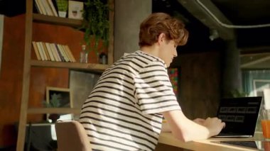 Modern dairede dizüstü bilgisayarda oturan gülümseyen adam. Genç bir adam yemek masasında otururken klavyede daktilo çalıyor. Yan atış. Modern iç tasarım. Kamera hareket ediyor