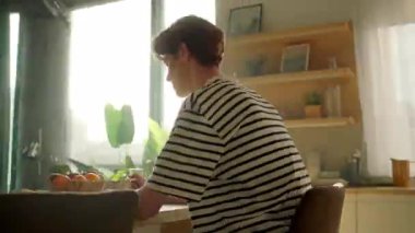 Enerjik hareketler, evden çalışan beyaz bir erkeğin görüntüsünü takip ediyor. Genç adam dizüstü bilgisayar kullanıyor ve mutfakta portakal suyu içiyor. Parallaks etkisi. Sinematik. 360 derece atış. Ev