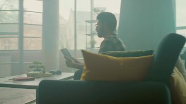 Evde gözlüklü genç adam koltukta oturan tablet bilgisayarı kullanıyor. Düşünüyorum. İletişim. Kamera hareket ediyor. 360 derece