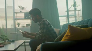 Gözlüklü mutlu genç adam evdeki koltukta oturup gülümseyen tablet bilgisayar kullanıyor. Düşünüyorum. İletişim. Kamera hareket ediyor. 360 derece