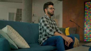 Gözlüklü şık genç adam kanepede dinleniyor. Akıllı telefon kullan. Sosyal medyada sohbet etmek. - Evde. Kamera hareket ediyor. 360 derece