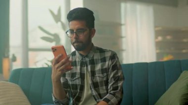 Kanepede dinlenen gözlüklü, gülümseyen genç yakışıklı adam akıllı telefon kullan. Sosyal medyada sohbet etmek. - Evde. Kamera hareket ediyor. 360 derece