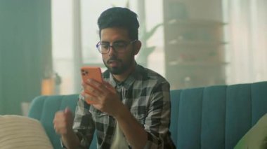 Kanepede dinlenen gözlüklü komik, gülümseyen genç adam akıllı telefon kullan. Sosyal medyada sohbet etmek. - Evde. Kamera hareket ediyor. 360 derece