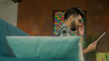 Gözlüklü sevimli sakallı adam evdeki kanepede oturan tablet bilgisayar kullanıyor. Gülümsemeyi düşünmek mutlu hissettiriyor. İletişim. Kamera hareket ediyor. 360 derece