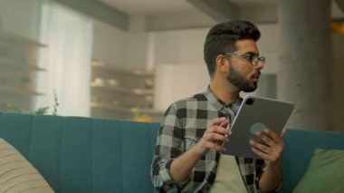 Gözlüklü komik sakallı adam evdeki kanepede oturan tablet bilgisayar kullanıyor. Gülümsemeyi düşünmek mutlu hissettiriyor. İletişim. Kamera hareket ediyor. 360 derece