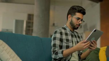 Gözlüklü mutlu sakallı adam evdeki koltukta otururken tablet bilgisayar kullanıyor. Düşünüyorum. İletişim. Kamera hareket ediyor. 360 derece