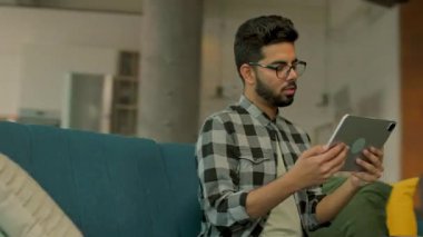 Gözlüklü, ciddi bir sakallı adam kanepeye oturmuş tablet bilgisayar kullanıyor. Düşünüyorum. İletişim. Kamera hareket ediyor. 360 derece