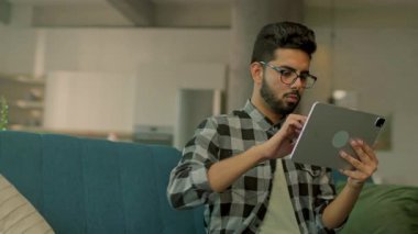 Modern evde, gözlüklü genç sakallı adam kanepeye oturmuş tablet bilgisayar kullanıyor. Düşünüyorum. İletişim. Kamera hareket ediyor. 360 derece