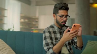 Kanepede dinlenen gözlüklü, mutlu genç adam ekranda akıllı telefon kullan. Sosyal medyada sohbet etmek. - Evde. Kamera hareket ediyor. 360 derece