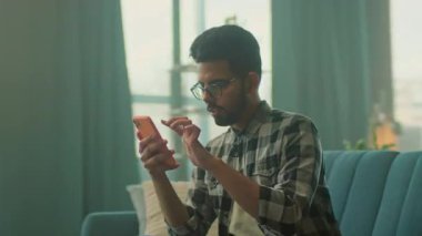 Kanepede dinlenen gözlüklü genç ve yakışıklı adama bakın. Akıllı telefon gülümsemesi kullanın. Sosyal medyada sohbet etmek. - Evde. Kamera hareket ediyor. 360 derece