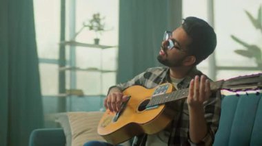 Genç adam gözlük takıyor ve evdeki oturma odasında kanepede gitar çalıyor. Gülümsüyor. Kamera hareket ediyor. 360 derece