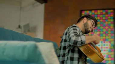 Gözlüklü genç adamı kapat ve evdeki oturma odasındaki kanepede gitar çal. Gülümsüyor. Kamera hareket ediyor. 360 derece