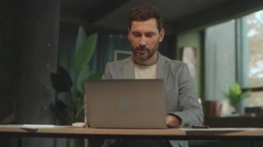 Masasında laptopuyla oturan mutlu beyaz adam başarıyı kutluyor. İş yerindeki heyecanlı stil sahibi iş adamı. İz sürme hareket ediyor. 360 derece atış. Hız. Sinematik