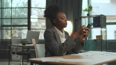 360 derecelik hızlandırma. Gri takım elbiseli Afro-Amerikalı iş kadını laptopuyla masada oturuyor ve akıllı telefon kullanıyor. Çalışan genç bir kadın. İz sürme sahnesinde hareketli enerji. Başarılı kız.
