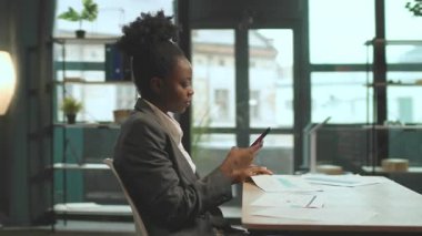 Masada otururken gülümseyen proje yöneticisi. Başarılı projeyi kutlayan çekici mutlu iş kadını. Genç Afro-Amerikan kadın akıllı telefon kullanıyor. 360 derece ofis. İz sürme