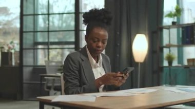 Düşünceli Afrikalı Amerikalı iş kadını masasında oturuyor ve ofisteki akıllı telefondan mesaj atıyor. Cep telefonuyla çalışan, müşterilerle sohbet eden odaklanmış genç bir kadın. 360 derecelik izleme atışı
