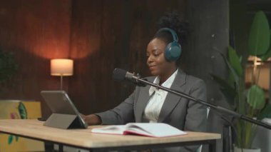 Güzel Afrika kökenli Amerikalı radyo sunucusu masasında oturmuş tablet ekranına dokunuyor ve podcast kaydına hazırlanıyor. Resmi giyinmiş yetenekli genç bir kadın. İş yeri konsepti. 360 derece