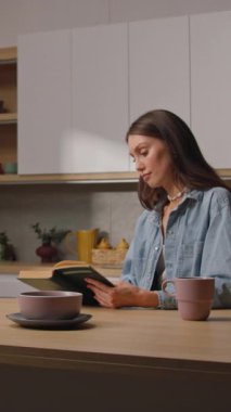 Çekici, kahverengi saçlı, rahat mutfak masasında oturmuş kitap okuyan genç bir kadının görüntüsü. Hafta sonu tatili. Boş zaman konsepti. Dışarıda yağmurlu bir gün