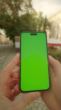 Şehir sokağının bulanık arka planında adamın ellerinin telefonu tutarken ve yeşil ekrana dokunurken yakın plan görüntüsü. Sosyal medyayı kontrol eden kişi. Krom anahtar. Mesajlaşma. Dikey pozisyonda göster. Açık havada.