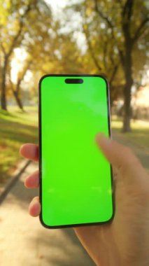 Kamu parkında yürürken cep telefonu kullanan genç beyaz bir adam. Yakın çekim erkek eli, yeşil ekranlı akıllı telefonu dikey pozisyonda tutuyor ve sosyal medyada dışarıya doğru kaydırıyor.