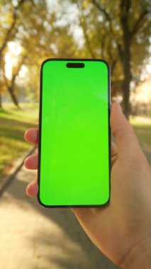 Sonbahar parkında yeşil ekran ve krom anahtarlı akıllı telefonu tutan genç bir erkeğin yakın görüntüsü. Telefon kullanan, sarı yapraklı ağaçların bulanık arka planında mesajlar okuyan bir adam.