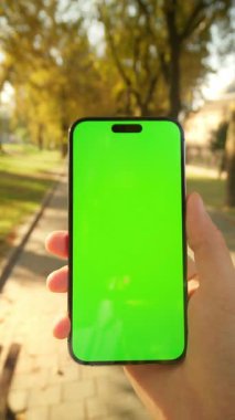 Sonbahar parkında elinde yeşil ekranlı cep telefonu olan bir gencin görüntüsü. Yakından erkek eli. Adam sokağın bulanık arka planından sosyal medyayı kontrol ediyor. Sarı yapraklı ağaçlar